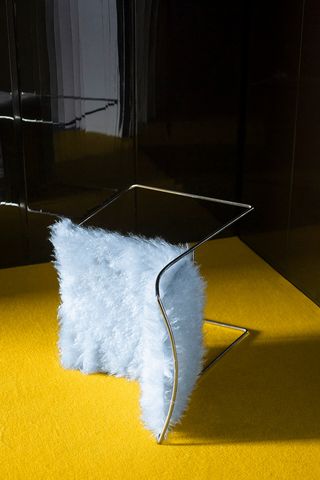 A retro-futuristic chair boasts a fluffy, light blue fur seating complete with a chrome base while sitting sideways on a bright yellow carpet.