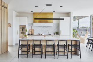 An open plan white kitchen with large island, black chairs and golden cooker hood