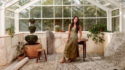 designer joanna gaines photographed in a green house orangery building with large glass windows and ceiling perched on a vintage stool and surrounded by the vintage style rugs from her latest designs with Magnolia Home by Joanna Gaines x Loloi