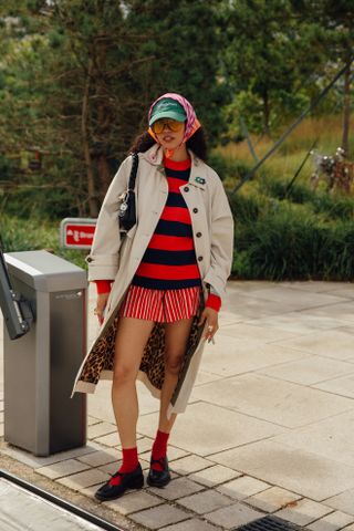 Woman at fashion week wearing preppy outfit featuring a red and blue striped sweater, red and white striped shorts, a tan trench coat, red socks, and black Mary Jane shoes.