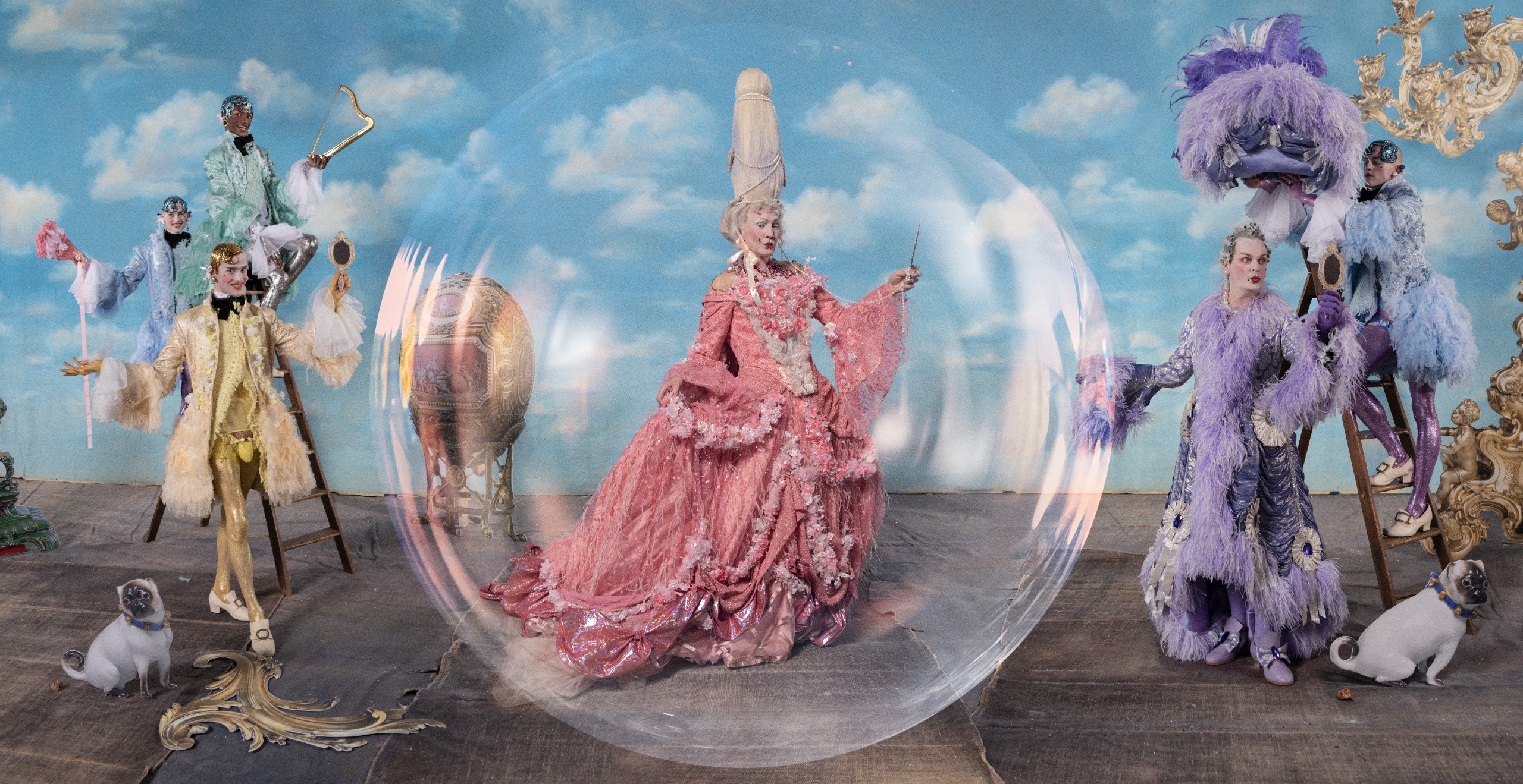 Photo of a rococo fantasy scene against a painted sky, with Paul O'Grady and Julian Clary in towering wigs and extravagant pink and lilac feathered gowns, one inside a giant bubble