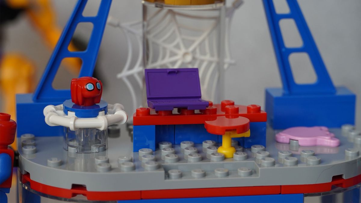 A small Spider-Man mask is on a platform next to a desk and chair with a web in the background, all in front of a grey background.