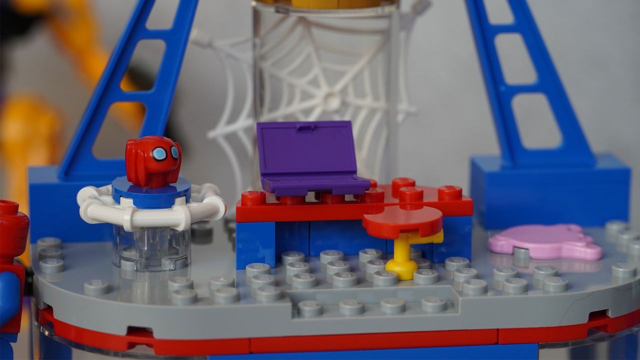 A small Spider-Man mask is on a platform next to a desk and chair with a web in the background, all in front of a grey background.
