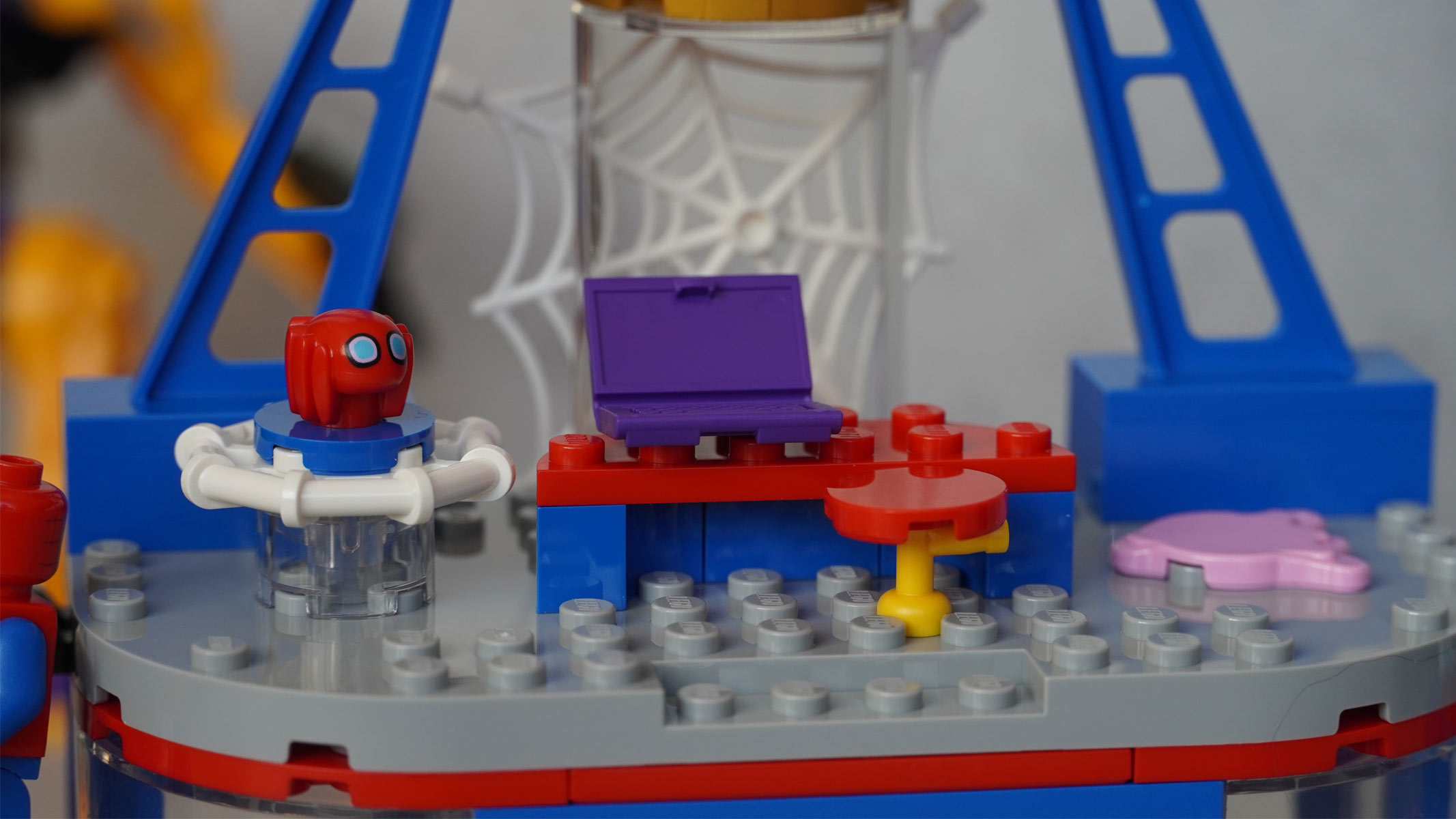 A small Spider-Man mask is on a platform next to a desk and chair with a web in the background, all in front of a grey background.