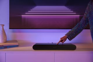 Sonos Ray i sort på en tv-bænk under et tv, hvor en person trykker på en af knapperne på toppen.