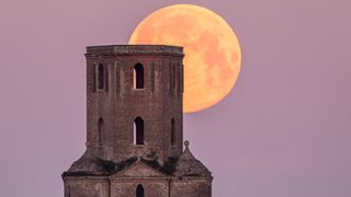 A large moon is rising behind an old stone tower against a pink hued sky.