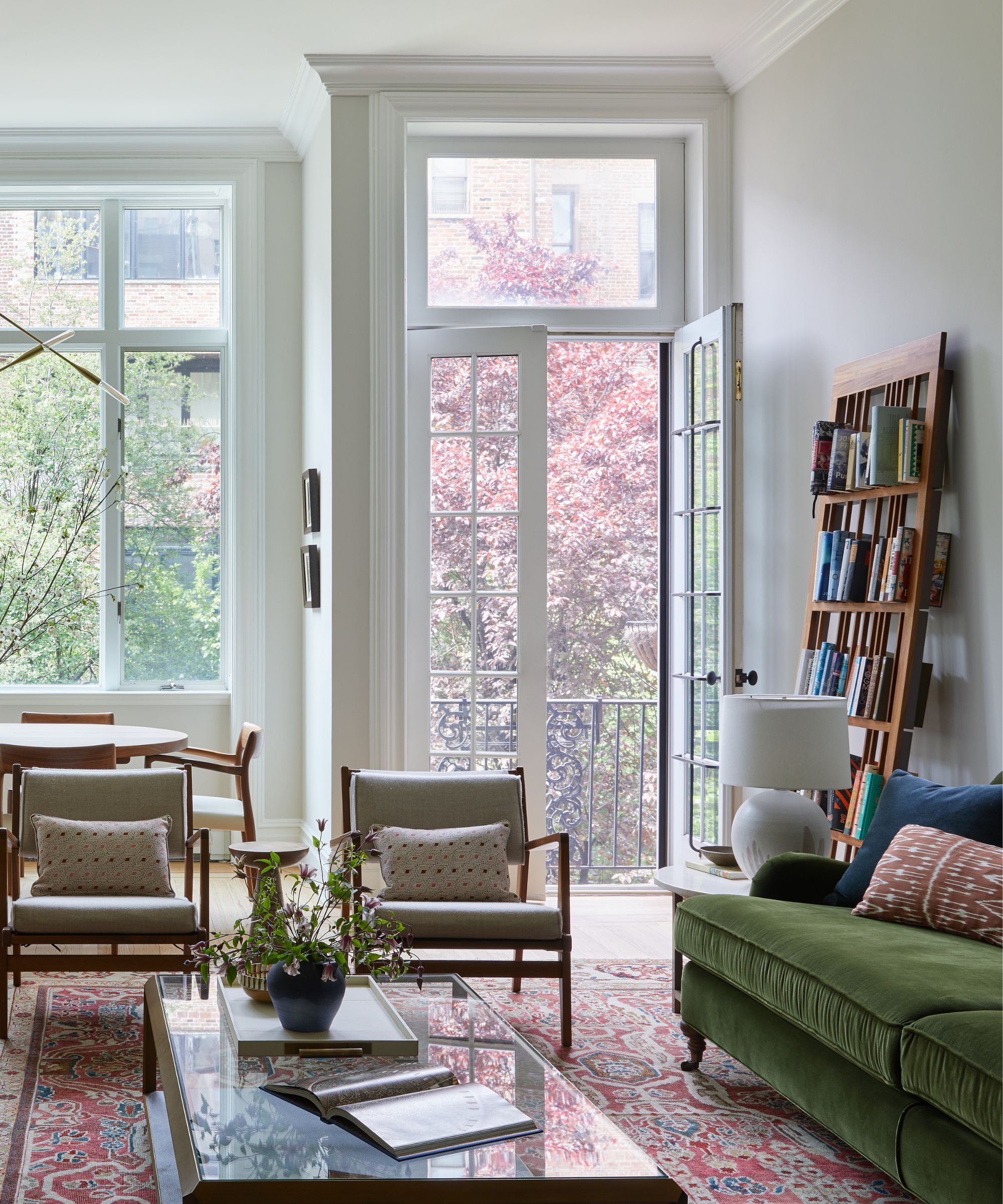 A transitional living room with light gray walls and high ceilings, a dark green sofa and two armchairs, and a red vintage rug. Balcony doors and large windows.