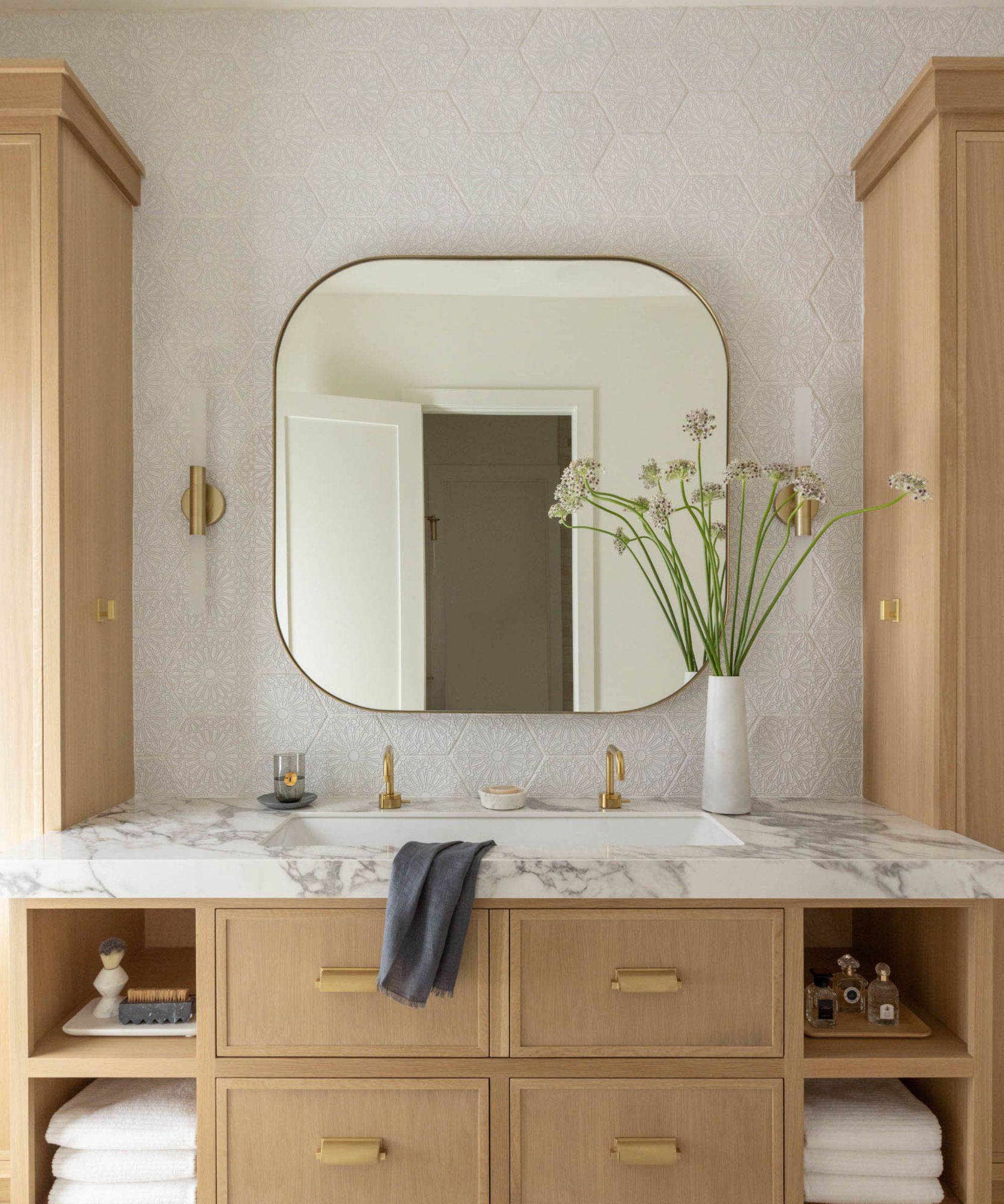 Neutral bathroom area with rounded square mirror, vase and flowers, small towel, marble countertop and white basin with gold faucets. Underneath are brown drawers and shelves.