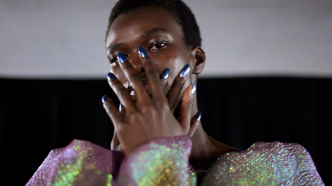 A model wearing an iridescent top holds a fresh manicure up to her face.