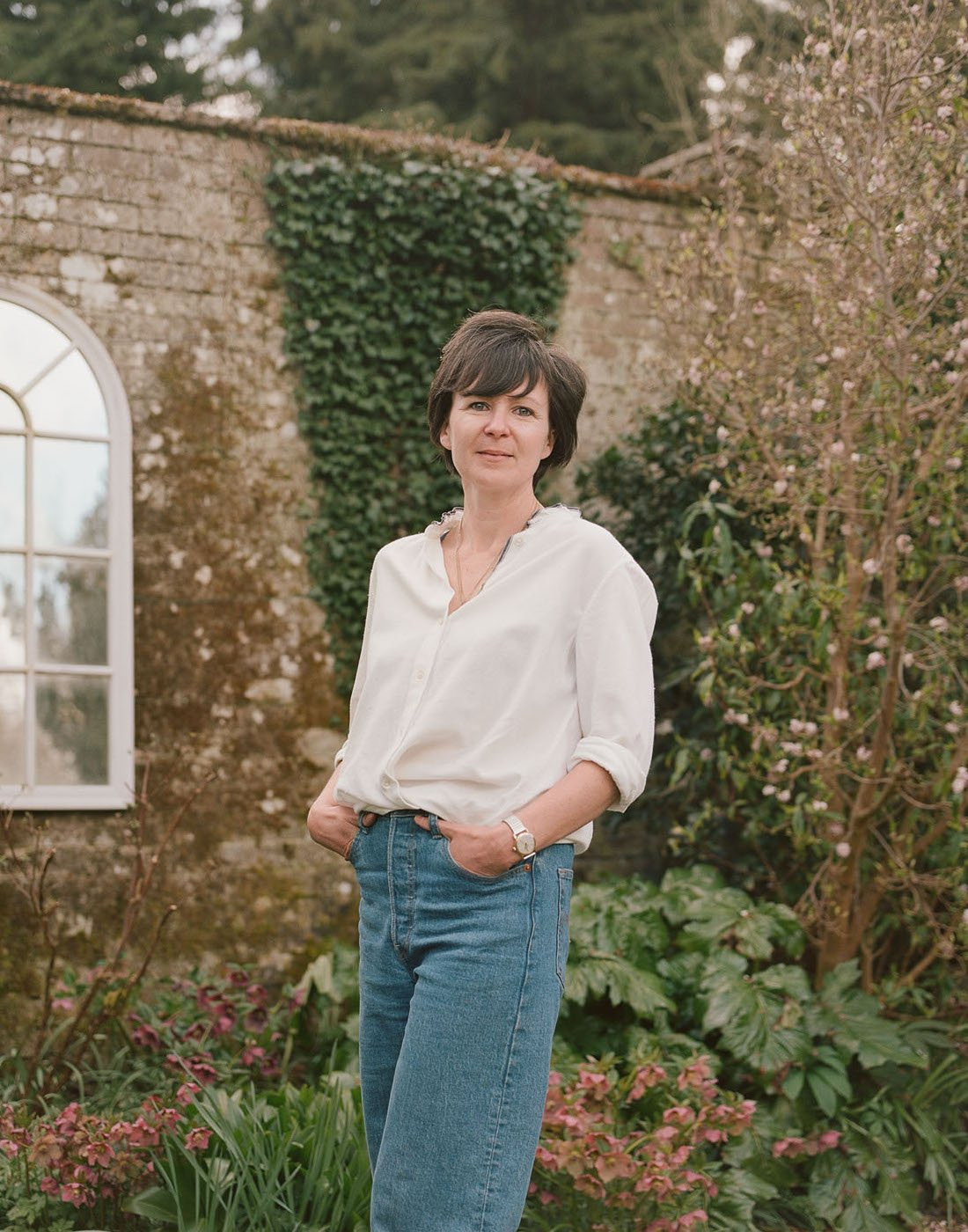 A woman with short dark hair cut in a straight bob wears a white shirt with folded up sleeves and a pair of loose jeans in front of a cottage wall covered in ivy and flowers.
