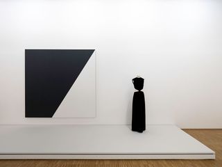 View of Centre Pompidou's tribute to Yves Saint Laurent featuring black and white wall art and one mannequin displaying a black dress in a space with white walls, a white platform and wood flooring