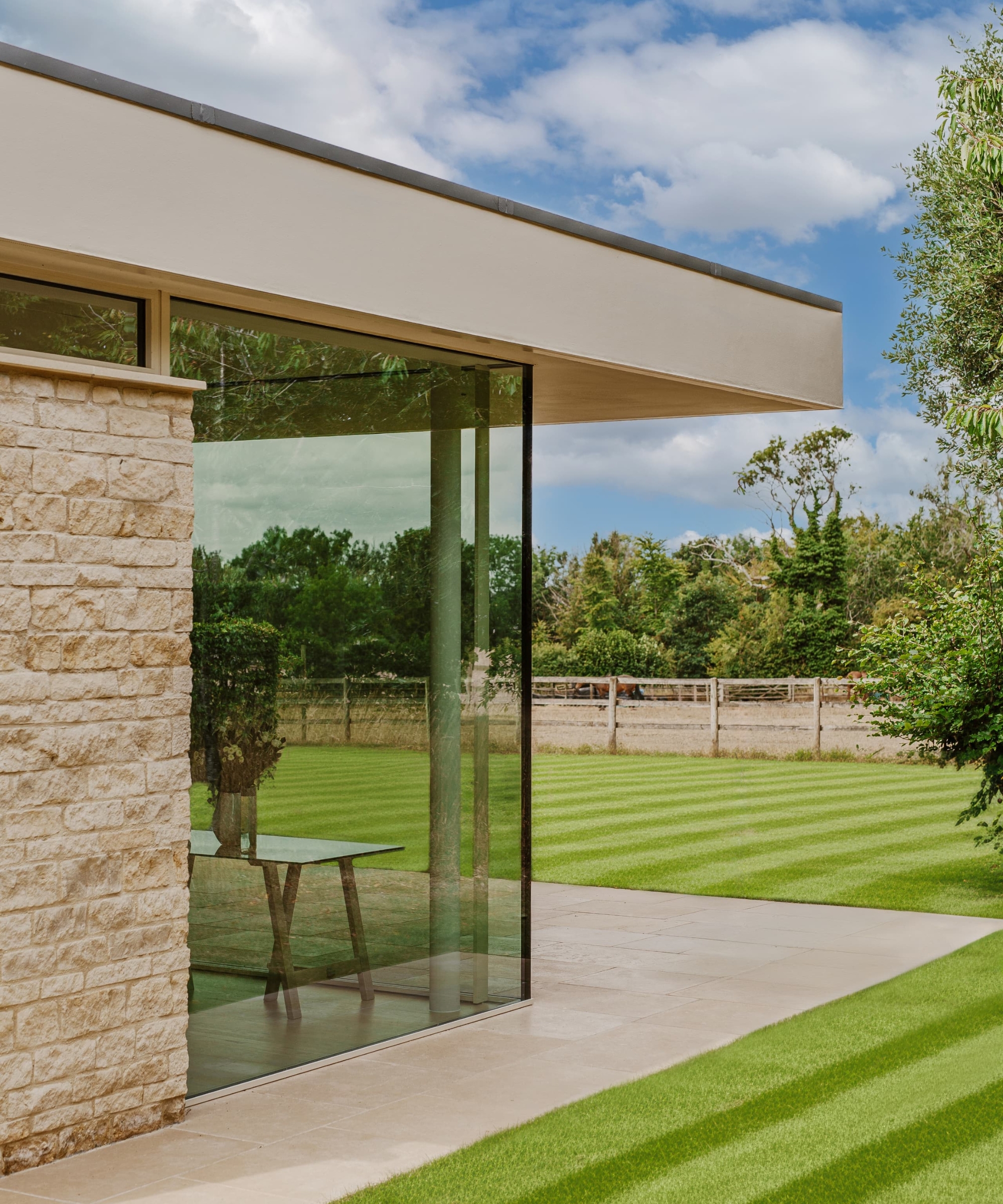 Frameless glass corner window in a stone-clad house opening onto a paved terrace and striped lawn.