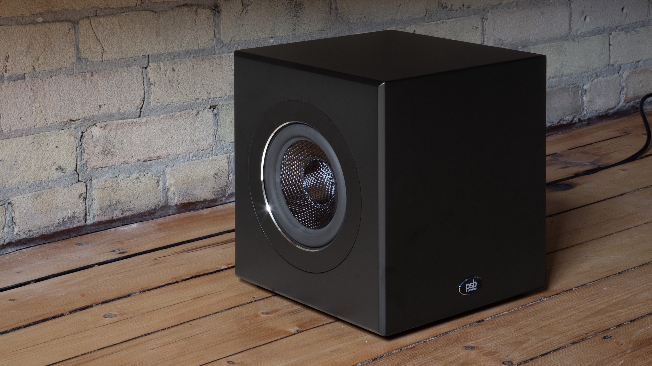 A PSB BP7 subwoofer on a wooden floor in front of a brick wall, shot at a 3/4 angle.
