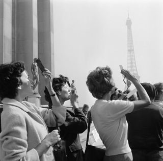 A photograph of three women amongst a crowd all looking up to the sky using smoked glass to view the eclipse. The Eiffel Tower is in the background.