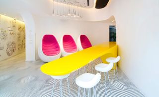 Interior view of The Poli House, Tel Aviv, Israel featuring white walls with illustrations, a white floor with a black wavy stripe design, pink and white chairs, a long yellow table and silver pendant lights