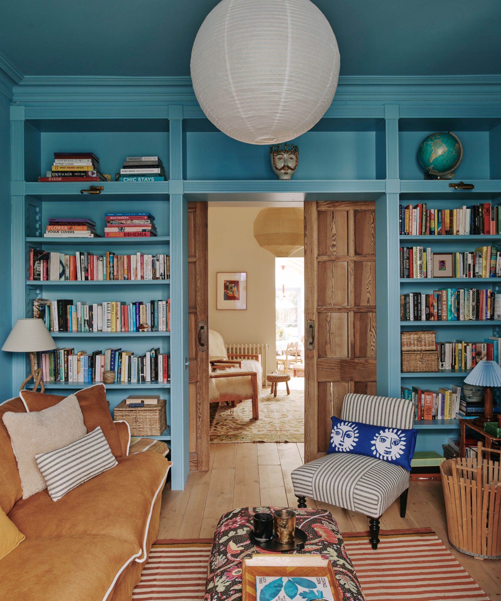 A cozy living room with blue color-drenched walls with built-in shelving, a brown sofa, a striped rug, and a patterned ottoman.
