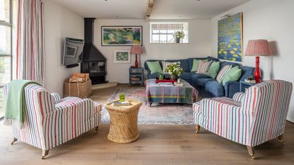 A bright and colorful living room with striped chairs, a patterned rug, blue corner sofa, rattan side table, black wood burner, and red lamps. The walls are white, with a large window to the left.