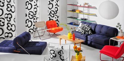 Image of an all-white room with IKEA retro furniture arranged around it. Three are three white curtains with a black doodle design, colorful modern shelving on the back wall, and a denim sofa and matching accent chair. A paper lantern hangs from the ceiling and there are white, orange, and red accent chairs as well.