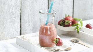 strawberry smoothie in a glass with a blue straw. The glass is on a white tray next to a bowl of strawberries