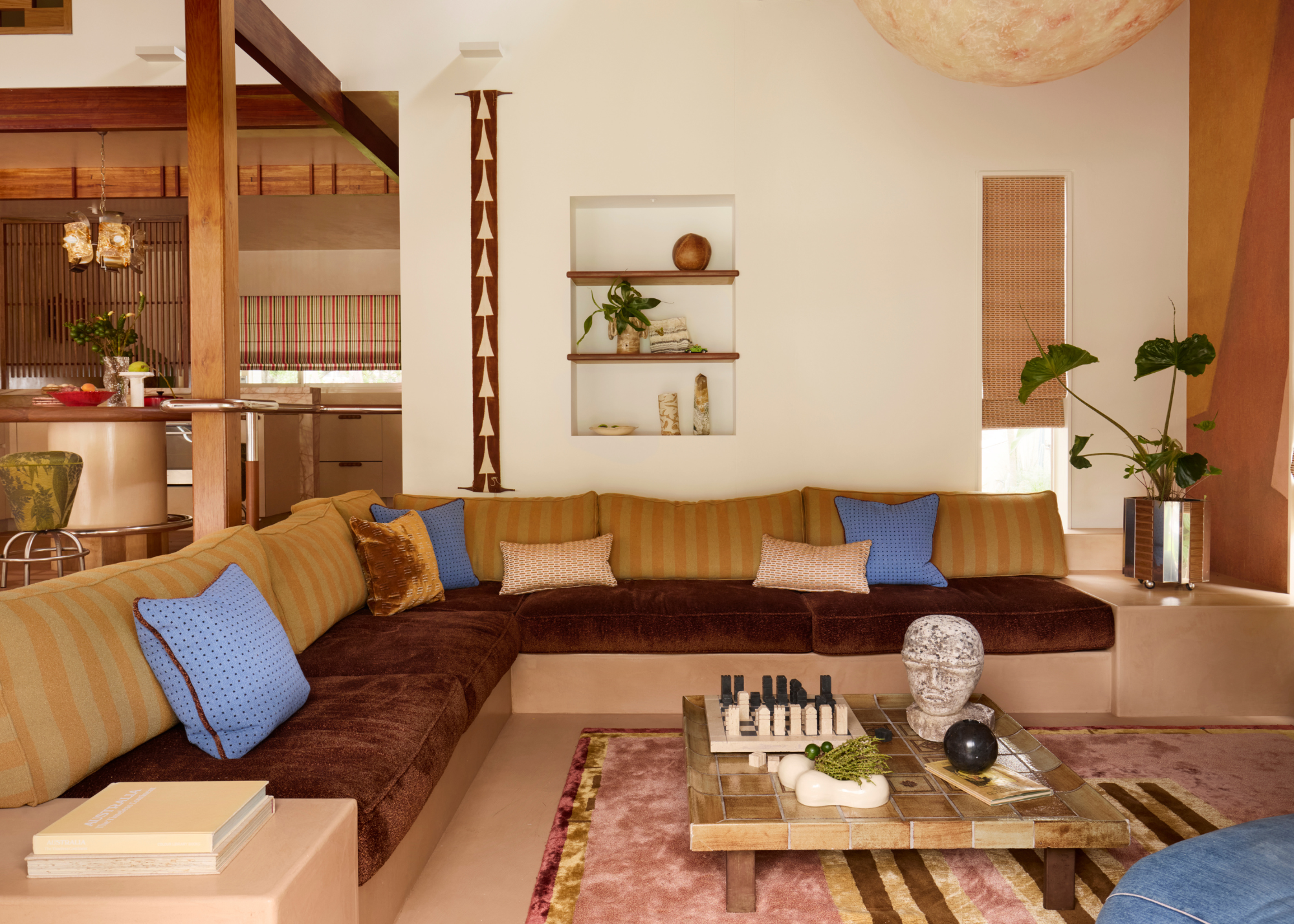 A built-in sunken, 70s-inpired lounge in earthy tones with pops of blue on the cushions, a low profile marble table with a black and cream stone chess set on top and a stone head sits underneath the large moon pendant light that anchors the space