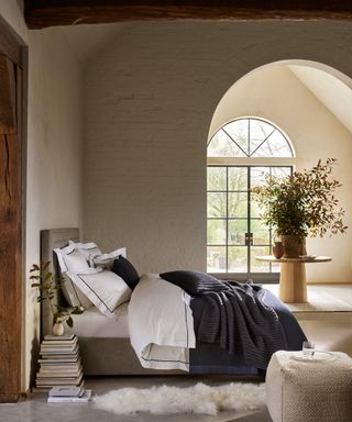 design a home that feels like you, bedroom with cream brick painted walls, large arched doorway to garden, beams, upholstered grey bed with blue and white bedding, table with foliage, books, sheepskin, footstool