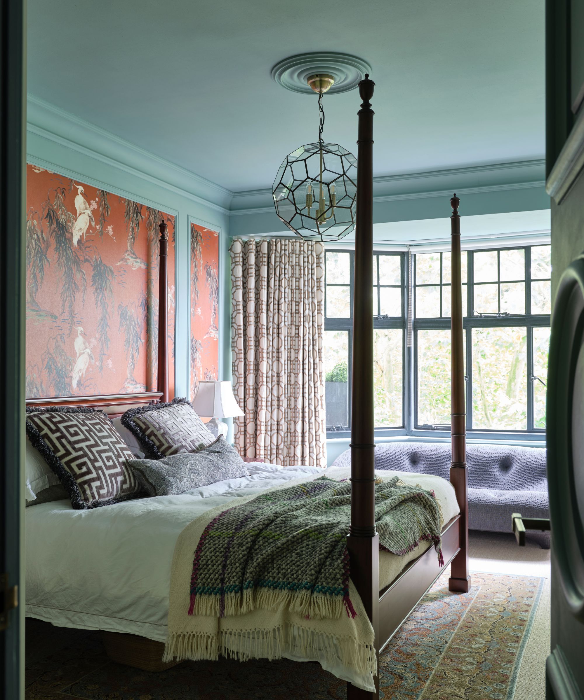 A blue bedroom with a four poster bed, orange wallpapered statement wall, patterned rug and large, bright bay windows.