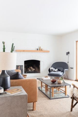 A living room by Modern Organic with white painted brick wall, grey armchair with footstool, coffee table, wooden shelf, and tan leather chair