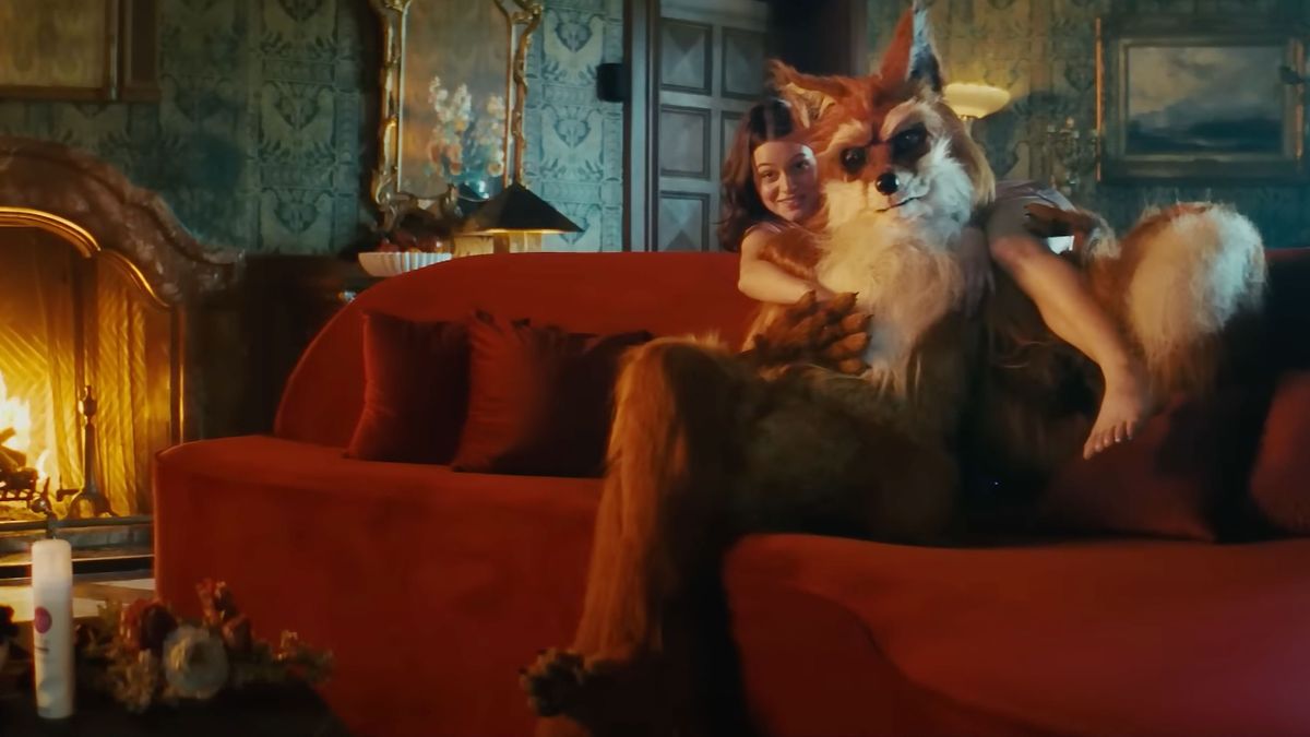 A shot of a humanoid fox in a dark room lounging on a sofa, with a human woman draped on its shoulders