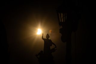 A partially eclipsed sun appears as a crescent shape and in the foreground is a statue with a figure holding up their hand.