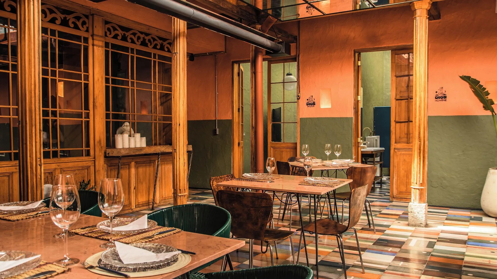 A terracotta-tinted indoor courtyard of a restaurant with colorful, geometric tiles, wooden furniture in industrial style matched with leather chairs, and tropical plants.