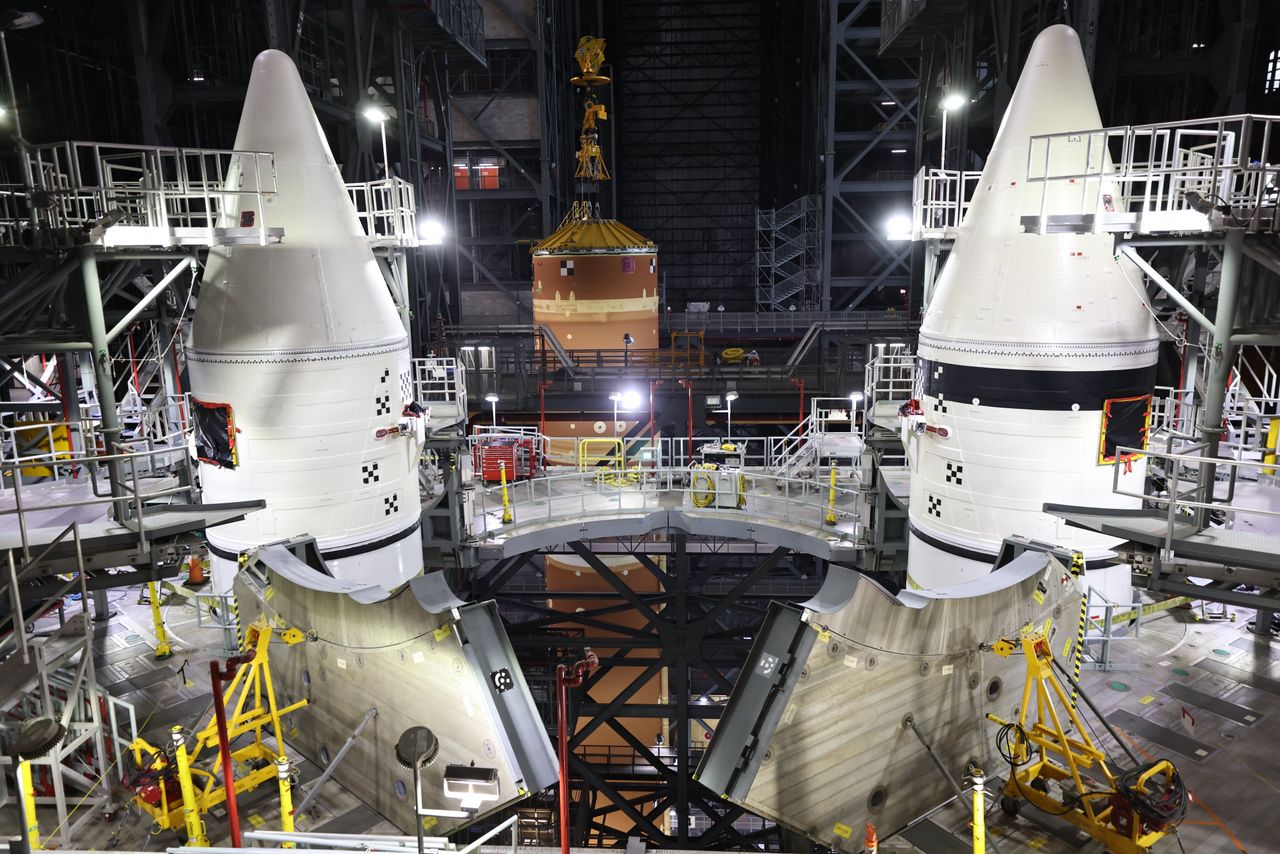 a large orange rocket booster is hoisted in a tall factory behind to giant pointy white boosters secured by scafolding.