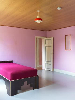 A bedroom with pink walls, a pink single bed with a wooden base, a wooden ceiling and a red pendant light.