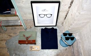 A downward view of an eye glasses poster on a wall with a step ladder, a suitcase, a folded blue shirt and sunglasses next to it.