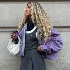 A woman wears a purple technical jacket with a grey pleated mini skirt and a white Miu Miu bag