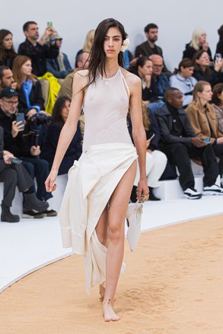 A female model wearing a short sleeve see through shirt and a long white dress walking down a runway made of beach sand.