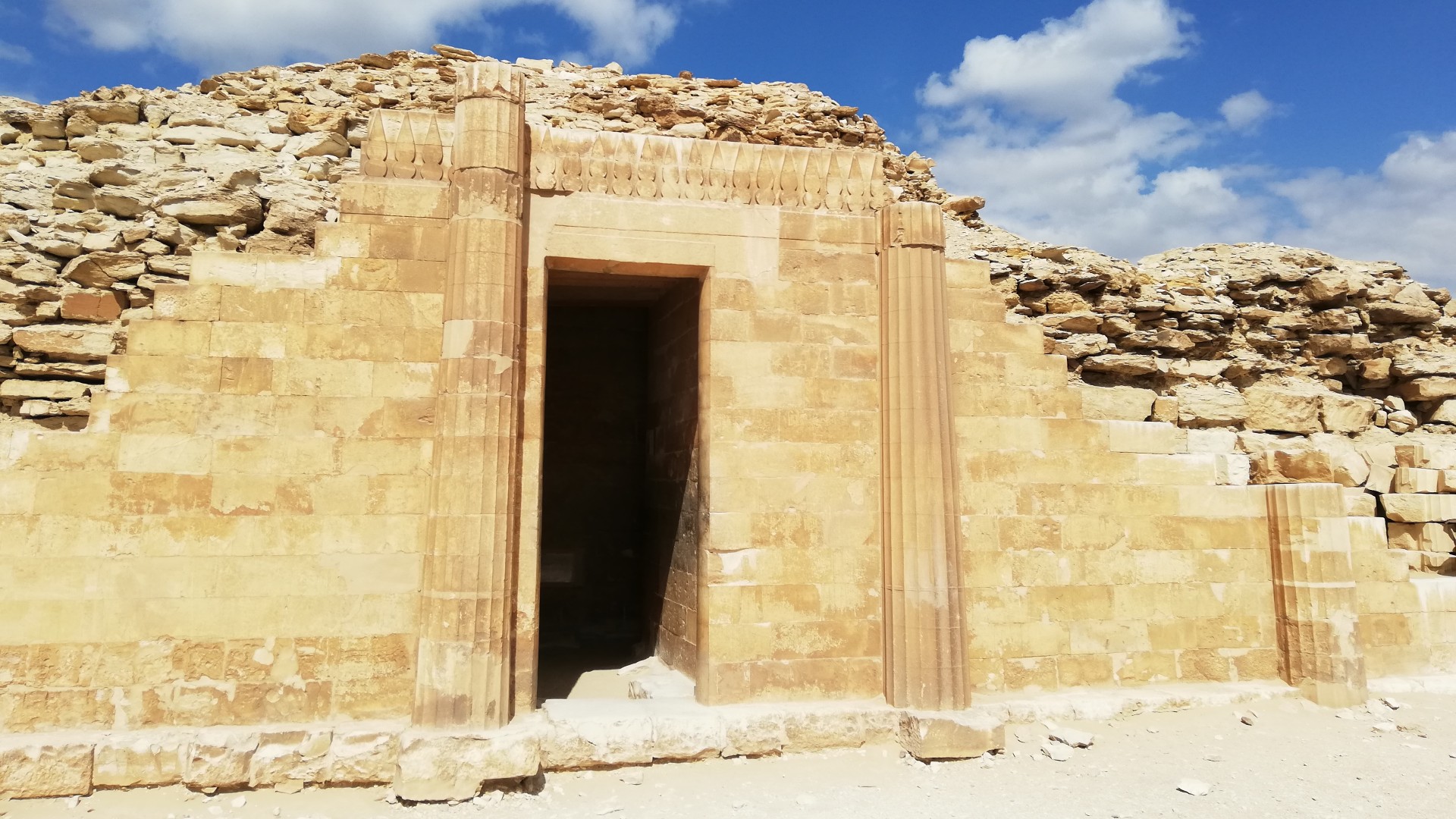 Here we see the pavilion at the funerary complex of pharaoh Djoser located in Saqqara, Egypt. We can see a doorway flanked by 2 tall columns. While there is mostly smooth bricks, at the top the bricks looked piled on more haphazardly and less neat.