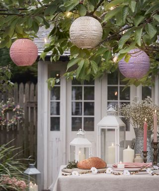 An outdoor space with pink, white, and purple lanterns in a green tree hanging over a pink dining table with white lamps, candles, and pink candlesticks, with a white gazebo behind it