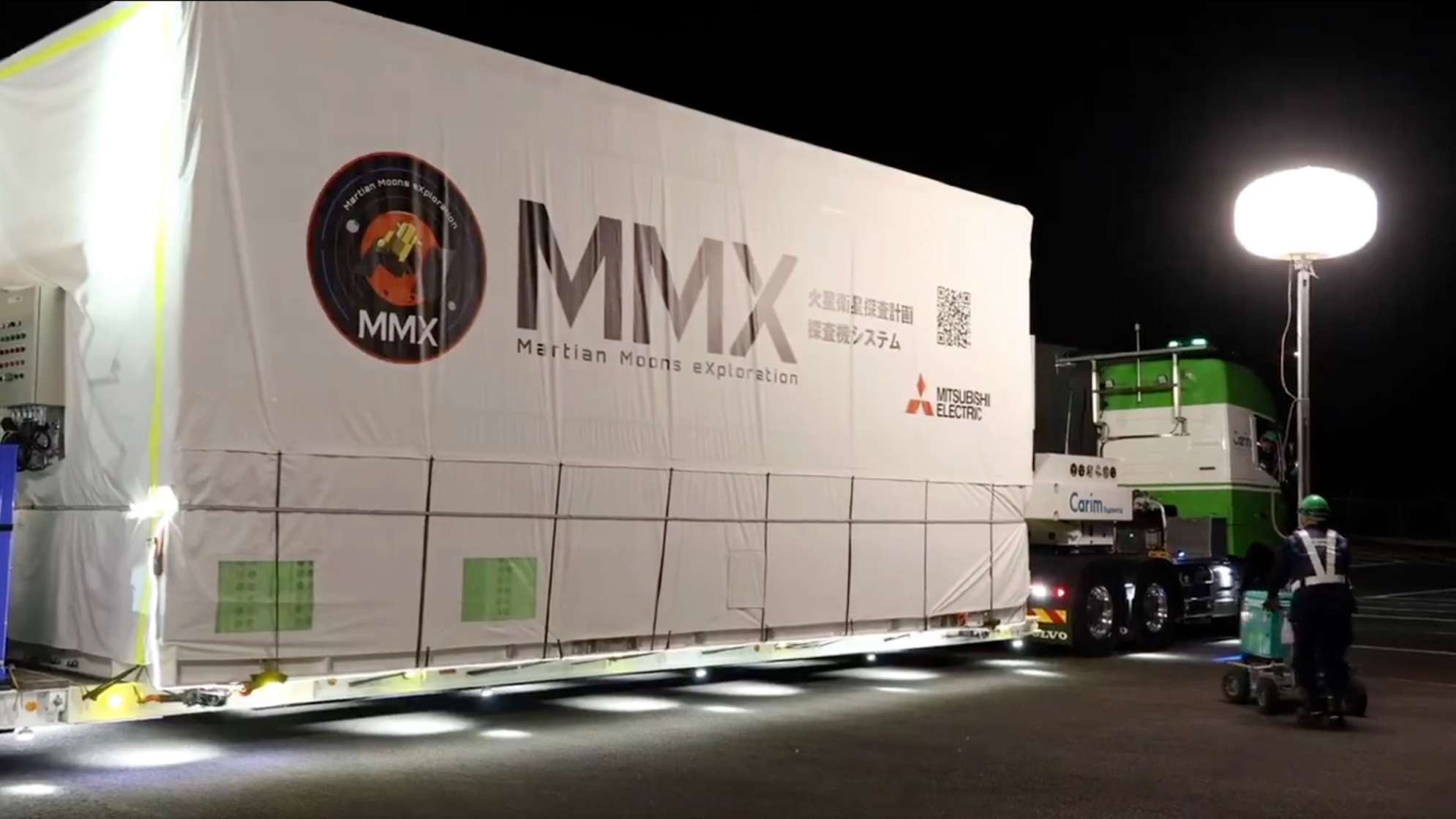 Closeup photo of a green semi truck delivering a large, rectangular white cargo payload with the words MMX on the side to its destination at night