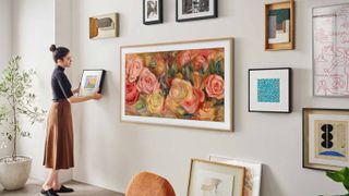 A woman hanging a small painting onto a wall alongside Samsung's The Frame TV, which is wrapped in wood-colored bezels and displaying an oil painting of flowers. Across the wall all several more pieces of artwork, emphasizing how The Frame blends into the space.