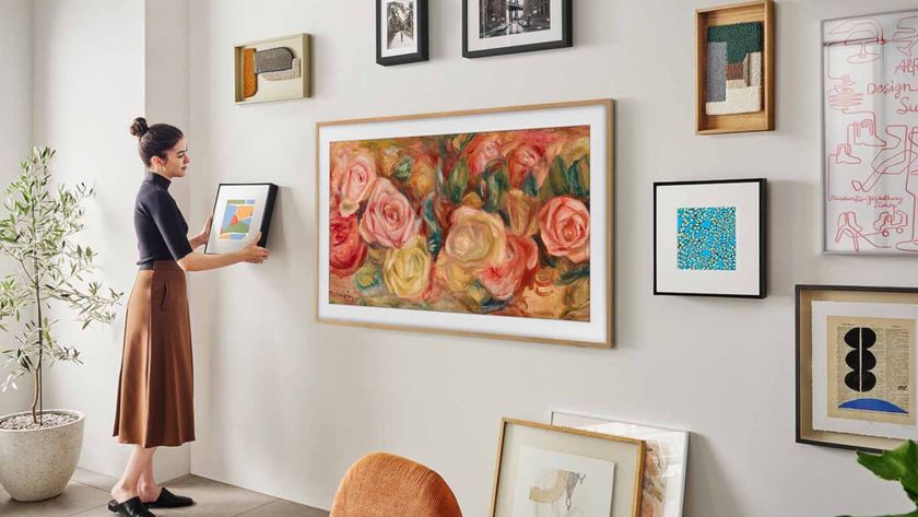 A woman hanging a small painting onto a wall alongside Samsung&#039;s The Frame TV, which is wrapped in wood-colored bezels and displaying an oil painting of flowers. Across the wall all several more pieces of artwork, emphasizing how The Frame blends into the space.