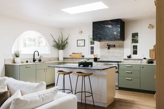 Short slab earthy colored backsplash in modern kitchen with black marble hood, island and sage green cabinet finish