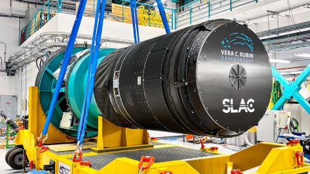 a massive black cylinder is lifted by crane into a round metal enclosure in a large warehouse