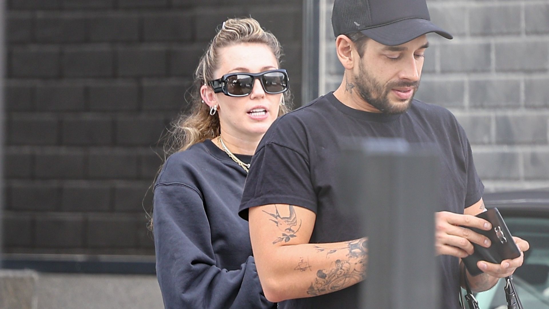 Miley Cyrus wears a black top and black track pants with green trainers and black sunglasses.