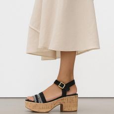 Woman wearing white skirt and black platform sandals with cork heels and white wall in background.