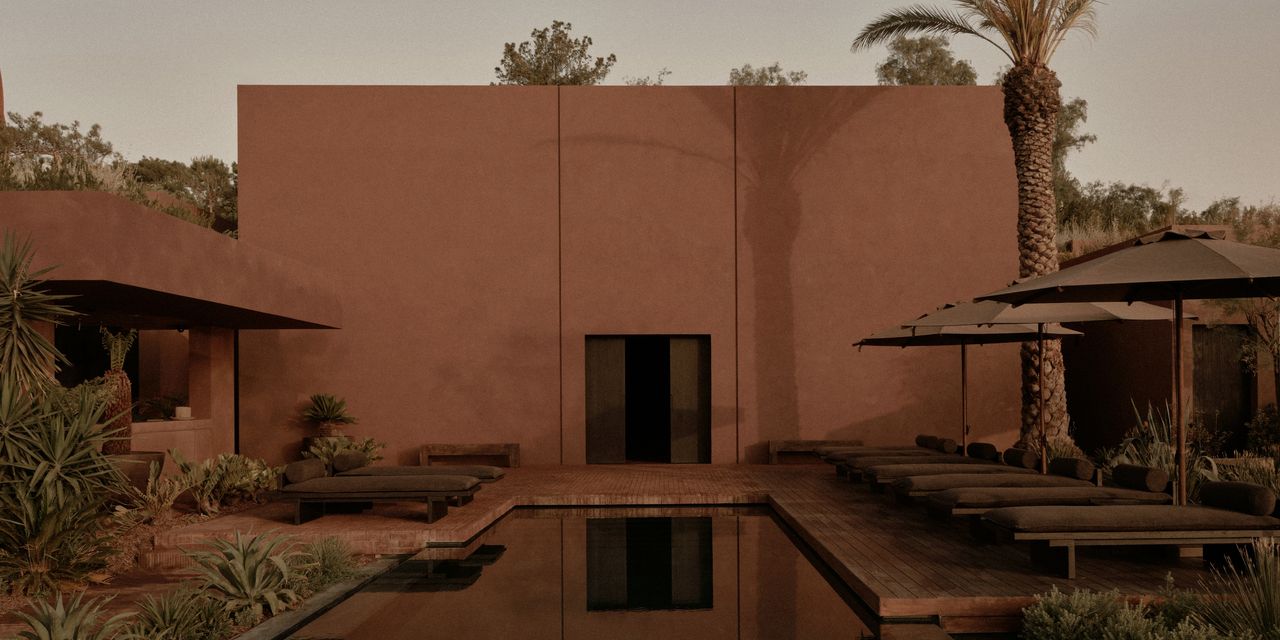 A terracotta-hued, adobe-style building is surrounded by similarly tinted organic furniture in wood, textile, and rattan and sprawling Mediterranean vegetation.