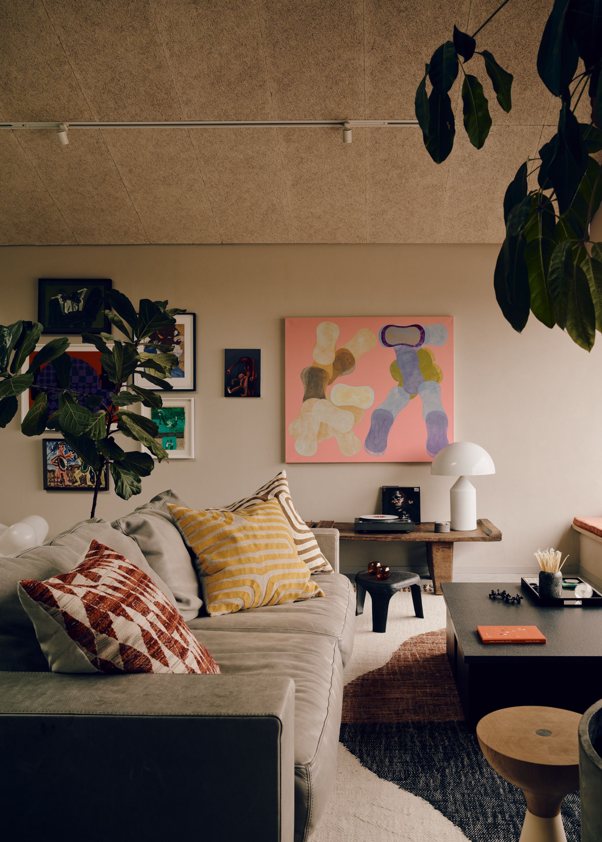 A modern living room with pale gray walls, a cork ceiling, a gallery wall with colorful artwork, a large gray low-slung sofa with yellow and red cushions, a black coffee table, and a large rug.