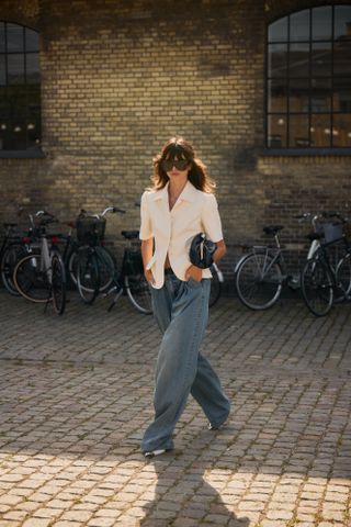 A woman wearing a tailored ivory top, baggy light-wash jeans, pointed toe shoes, oversized sunglasses, and a large black soft clutch.