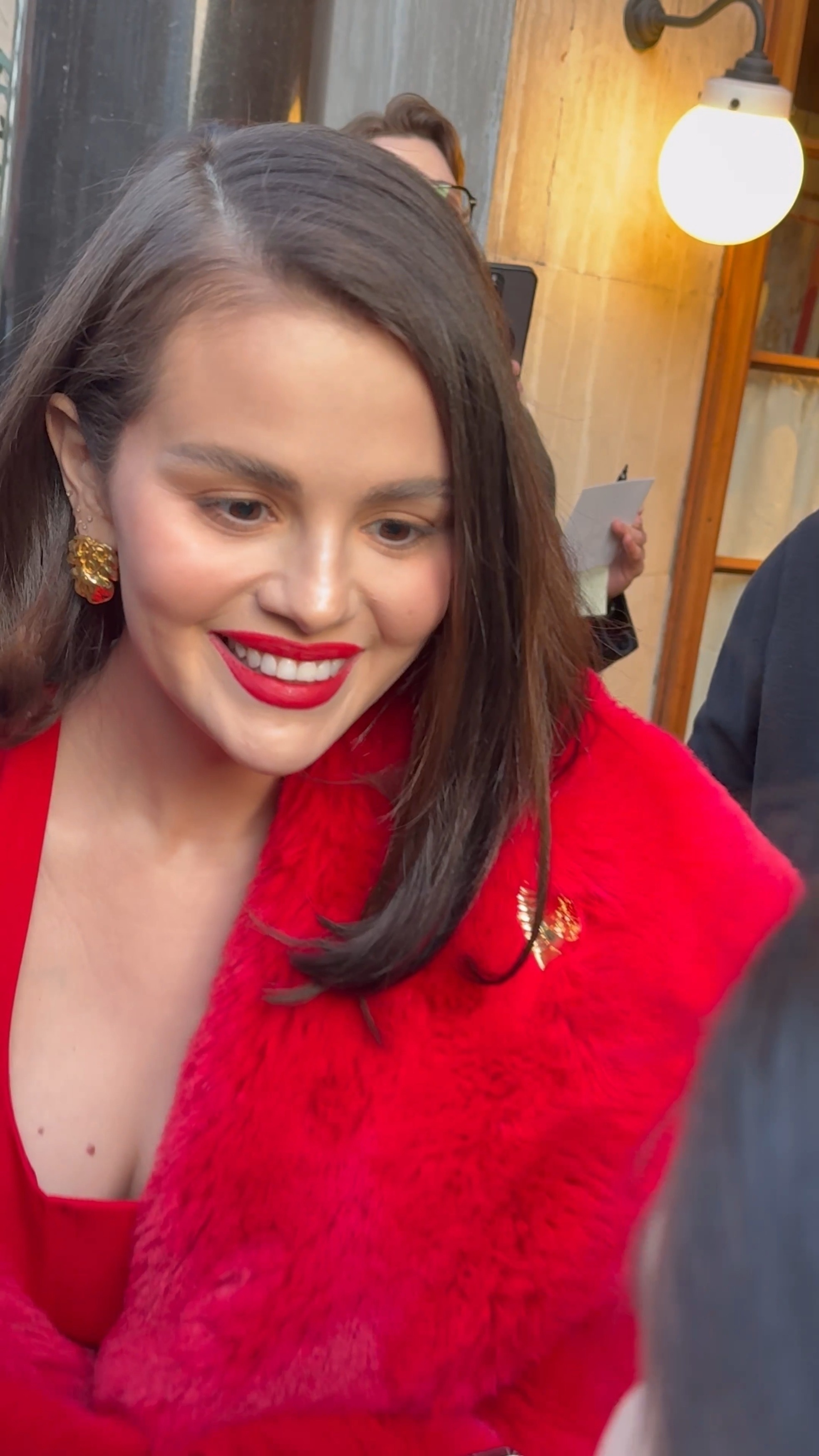 Selena Gomez in London wearing a red faux fur coat with a gold brooch and matching lipstick