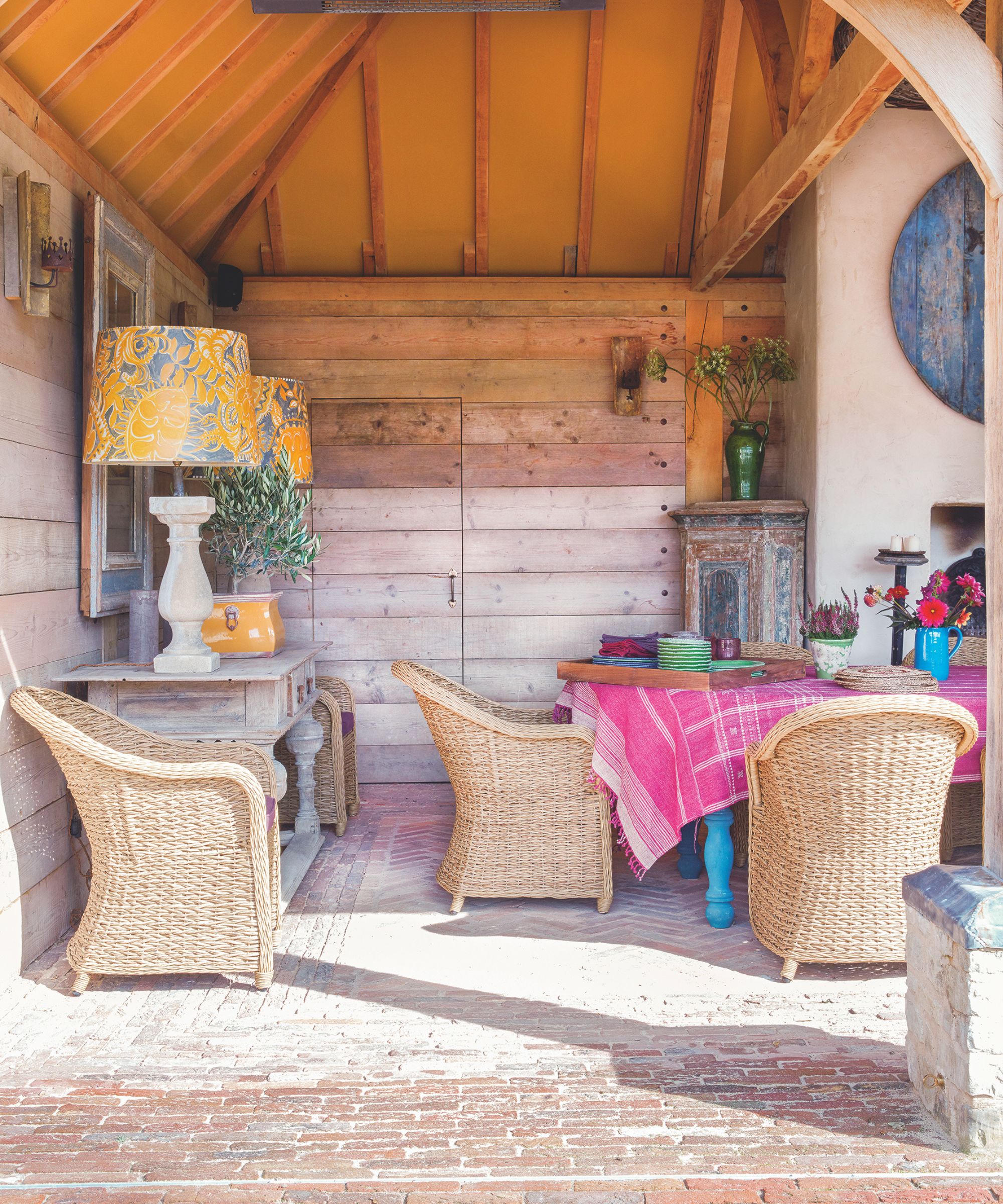 Outdoor barn conversion with brick floor, rattan armchairs dotted around a blue dining table with a pink tablecloth, and two large yellow table lamps on a console.