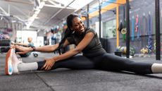 A woman sits on the floor of her gym with her straight legs out wide. She leans towards her right leg and holds the top of her right sneaker in her hand.